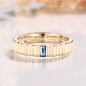 May include: A gold-colored ring with a vertical ridged design. The ring features a rectangular, dark blue gemstone set in the center. The ring is displayed on a reflective surface.