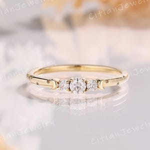 May include: A delicate gold ring featuring three clear, round gemstones set in a row. The band is adorned with small stars and crescent moon designs. The ring is set against a soft, blurred background.