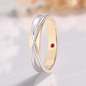 May include: A two-tone ring with a gold interior and a silver overlay that crosses over the front. A small, round, red gemstone is set into the gold band. The ring is a piece of jewelry.