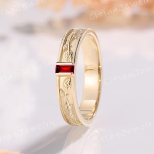 Könnte beinhalten: Ein goldener Bandring mit einem rechteckigen, im Baguetteschliff geschliffenen roten Edelstein, der horizontal eingesetzt ist. Die Oberfläche des Metallrings ist mit einem durchgehenden Muster aus zarten Blättern und Ranken graviert.