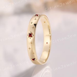 Peut inclure: Une bague dorée avec une finition polie. La bague est ornée de gravures en forme d'étoiles et de croix, ainsi que de petites pierres précieuses rouges. Il s'agit d'un bijou.