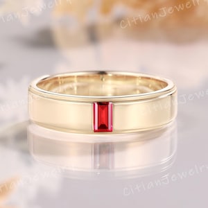 May include: A gold-colored ring with a rectangular, vibrant red gemstone set in the center. The ring has a flat, polished band and a simple, elegant design, perfect for a special occasion.
