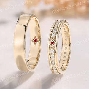May include: Two gold wedding rings. One is a simple band with a small red gemstone. The other ring is encrusted with small diamonds and a red gemstone. The rings are displayed on a white surface.