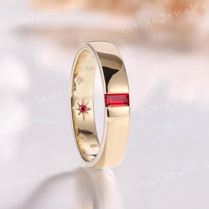 May include: A gold ring featuring a rectangular, red gemstone. The ring has a flat band and a small star and moon design on the inner side. The ring is displayed on a reflective surface. The ring is a piece of jewellery.