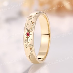 Puede incluir: Un anillo de banda de oro con acabado pulido y un patrón celestial grabado de estrellas y un motivo de rayos de sol. Una pequeña piedra preciosa roja y redonda está engastada en el centro del diseño solar. El texto CitianJewelry es visible.