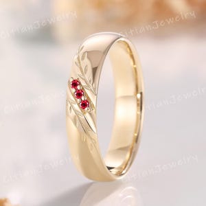 Puede incluir: Un anillo de banda de oro pulido con tres pequeños rubíes rojos redondos engastados en una fila vertical. La superficie de la banda de oro está decorada con delicados patrones de hojas grabados que se extienden a lo largo del anillo.