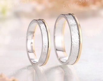 May include: Two wedding rings with a silver band and a textured, hammered silver center. The rings have a gold-colored edge. The background is blurred with soft, neutral tones. The rings are displayed on a reflective surface.