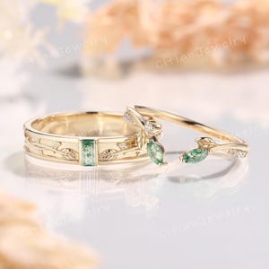 May include: Two gold wedding rings. One ring features a rectangular green gemstone and leaf design. The other ring has two green leaf-shaped gemstones and small white gemstones. The rings are on a white surface.
