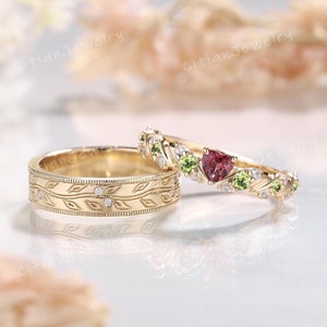 Könnte beinhalten: Zwei goldene Eheringe. Ein Ring hat ein Blattmuster mit kleinen Diamanten. Der andere Ring hat einen herzförmigen roten Edelstein, grüne Edelsteine und kleine Diamanten. Die Ringe liegen auf einer reflektierenden Oberfläche.
