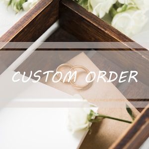 May include: Two gold wedding bands in a wooden box with a white card that says "CUSTOM ORDER" and a white rose.