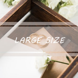 May include: Two gold wedding bands in a wooden box with a white card and a white rose. The card says "LARGE SIZE".