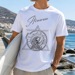 Puede incluir: Camiseta blanca con la palabra "Acuario" en escritura cursiva sobre un diseño geométrico con símbolos del zodiaco y una criatura marina. El diseño es en blanco y negro. La camiseta es de manga corta.
