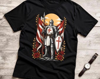 May include: A black t-shirt featuring a graphic print of a knight in silver armor holding a polearm and a shield with a red cross. The design includes red roses, striped flags, and a yellow sun background. A wristwatch is placed next to the shirt.