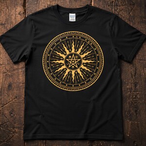 Puede incluir: Una camiseta negra de manga corta con un gran gráfico circular dorado en el pecho. El diseño incluye un pentagrama central rodeado de rayos solares, flechas, pequeñas estrellas y lunas crecientes dentro de un borde circular.