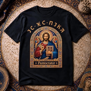 Puede incluir: Una camiseta negra de manga corta con un gráfico central de una figura barbuda con túnicas azules y rojas que sostiene un libro. Arriba hay texto dorado estilizado y debajo la palabra "Pantocrator" enmarcada por un arco dorado ornamentado.