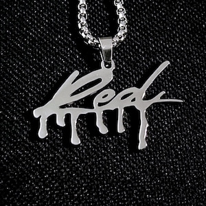 Könnte beinhalten: Halskette mit silberfarbenem Anhänger mit dem Wort "Red" in einer stilisierten, tropfenden Schrift. Der Anhänger hängt an einer silbernen Kette. Der Hintergrund ist ein dunkler, strukturierter Stoff.