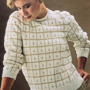 Op de afbeelding: Crèmekleurige trui met een rasterpatroon in contrasterend goud. De trui heeft een ronde hals en lange mouwen met geribde manchetten. Het rasterontwerp bevat kleine ruitvormige uitsparingen, wat een delicate touch toevoegt aan de knitwear.