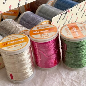 May include: A selection of WonderFil rayon thread spools in shades like metallic silver, vibrant pink, and green. The spools are labelled with the brand name and thread type, including "Razzle" and "Dazzle". The thread is wound on clear plastic spools.