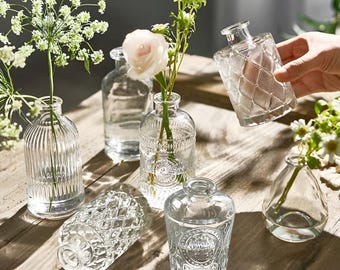 May include: Six clear glass vases of varying shapes and sizes, with textured surfaces, are arranged on a wooden table. Some vases are filled with white and pink flowers.