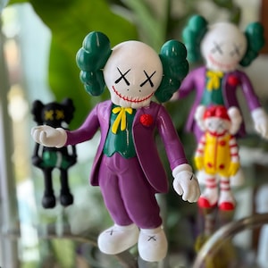 May include: A collection of colorful figurines, including a white figure with green ears, a purple suit, and a red flower. Other figures include a black figure and a figure in a yellow and red outfit. The figures have white gloves and feet.