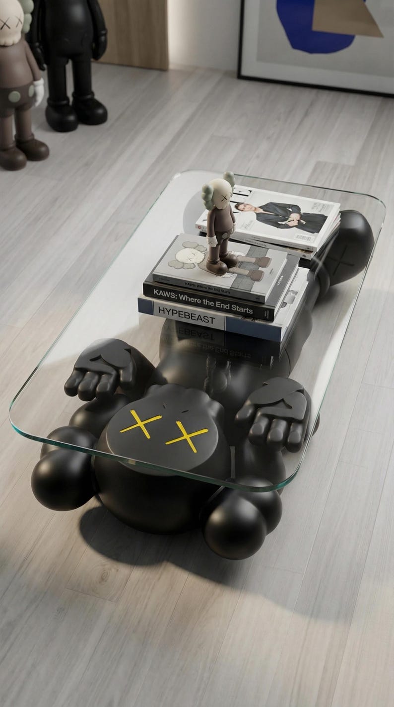 Pode incluir: Uma mesa de centro preta em forma de personagem de desenho animado com tampo de vidro. A figura tem ossos cruzados amarelos no rosto. Livros e uma estatueta est&atilde;o na superf&iacute;cie de vidro.