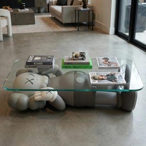 Pode incluir: Uma sala de estar moderna apresenta uma mesa de centro &uacute;nica. A mesa tem um tampo de vidro transparente suportado por uma figura cinza de desenho animado com luvas brancas e olhos riscados. Livros e revistas s&atilde;o exibidos na mesa.