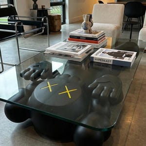 Pode incluir: Uma mesa de centro moderna com um tampo de vidro transparente e uma base escultural preta. A base &eacute; uma figura estilizada com olhos amarelos em forma de cruz. Livros empilhados na mesa.