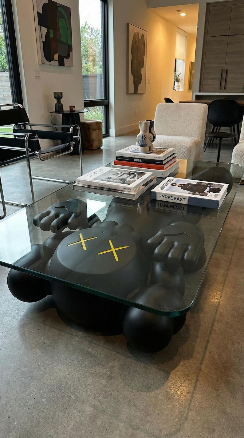Pode incluir: Uma mesa de centro moderna com um tampo de vidro transparente e uma base escultural preta. A base &eacute; uma figura estilizada com olhos amarelos em forma de cruz. Livros empilhados na mesa.
