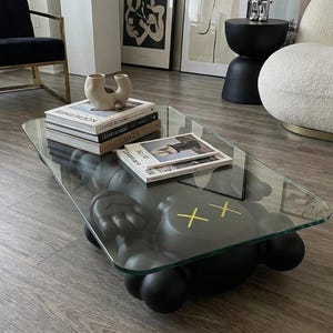 Pode incluir: Uma mesa de centro moderna com um tampo de vidro transparente e uma base cinza escuro. A base apresenta uma figura estilizada com olhos de cruz amarelos. H&aacute; livros e revistas na mesa, e a sala tem obras de arte e objetos decorativos.