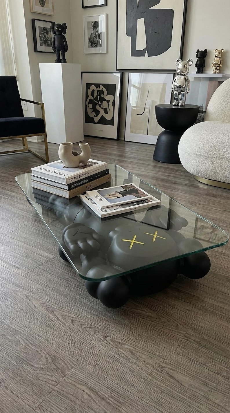 Pode incluir: Uma mesa de centro moderna com um tampo de vidro transparente e uma base cinza escuro. A base apresenta uma figura estilizada com olhos de cruz amarelos. H&aacute; livros e revistas na mesa, e a sala tem obras de arte e objetos decorativos.