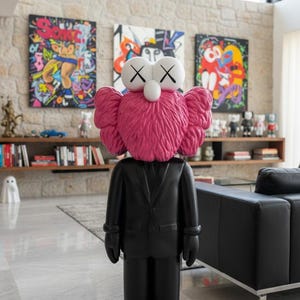 May include: A large black sculpture of a figure in a suit with a pink, furry head and white eyes with black X's. The sculpture is standing in a modern living room with colorful artwork and a black leather sofa.