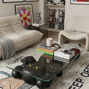 Pode incluir: Uma sala de estar moderna com uma mesa de centro preta com base de figura KAWS e tampo de vidro. O quarto inclui obras de arte, figuras decorativas, um tapete com texto e um sof&aacute; branco.