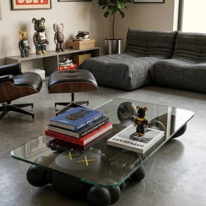 Pode incluir: Uma sala de estar moderna com uma mesa de centro com tampo de vidro e base preta com bordas arredondadas. A mesa cont&eacute;m livros e uma pequena figura. Obras de arte e um sof&aacute; cinza est&atilde;o ao fundo, junto com figuras decorativas.