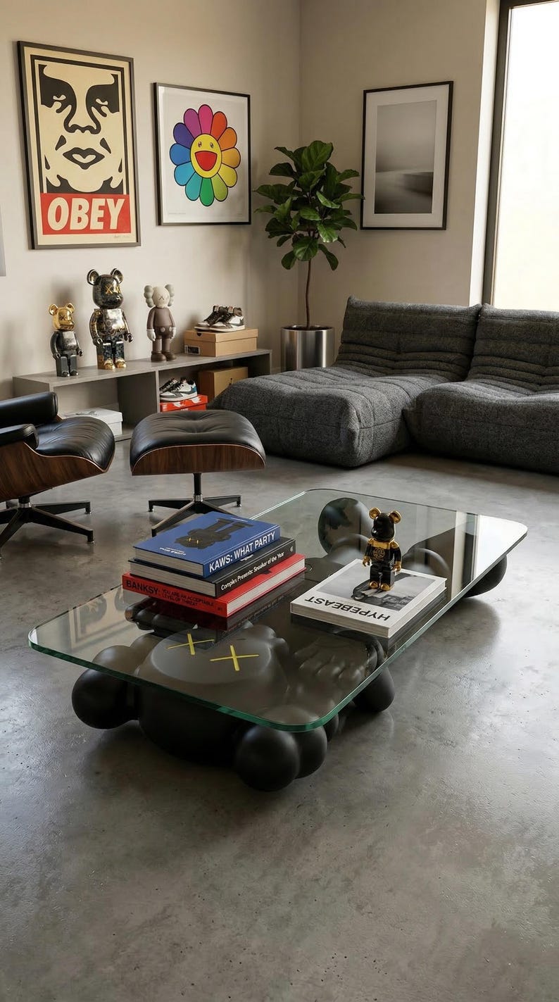 Pode incluir: Uma sala de estar moderna com uma mesa de centro com tampo de vidro e base preta com bordas arredondadas. A mesa cont&eacute;m livros e uma pequena figura. Obras de arte e um sof&aacute; cinza est&atilde;o ao fundo, junto com figuras decorativas.