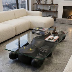 Pode incluir: Uma sala de estar moderna com uma mesa de centro preta com design KAWS com tampo de vidro. A mesa tem um design de figura &uacute;nico com olhos amarelos em forma de cruz. Livros e itens decorativos s&atilde;o colocados na mesa. Um sof&aacute; branco e obras de arte est&atilde;o ao fundo.