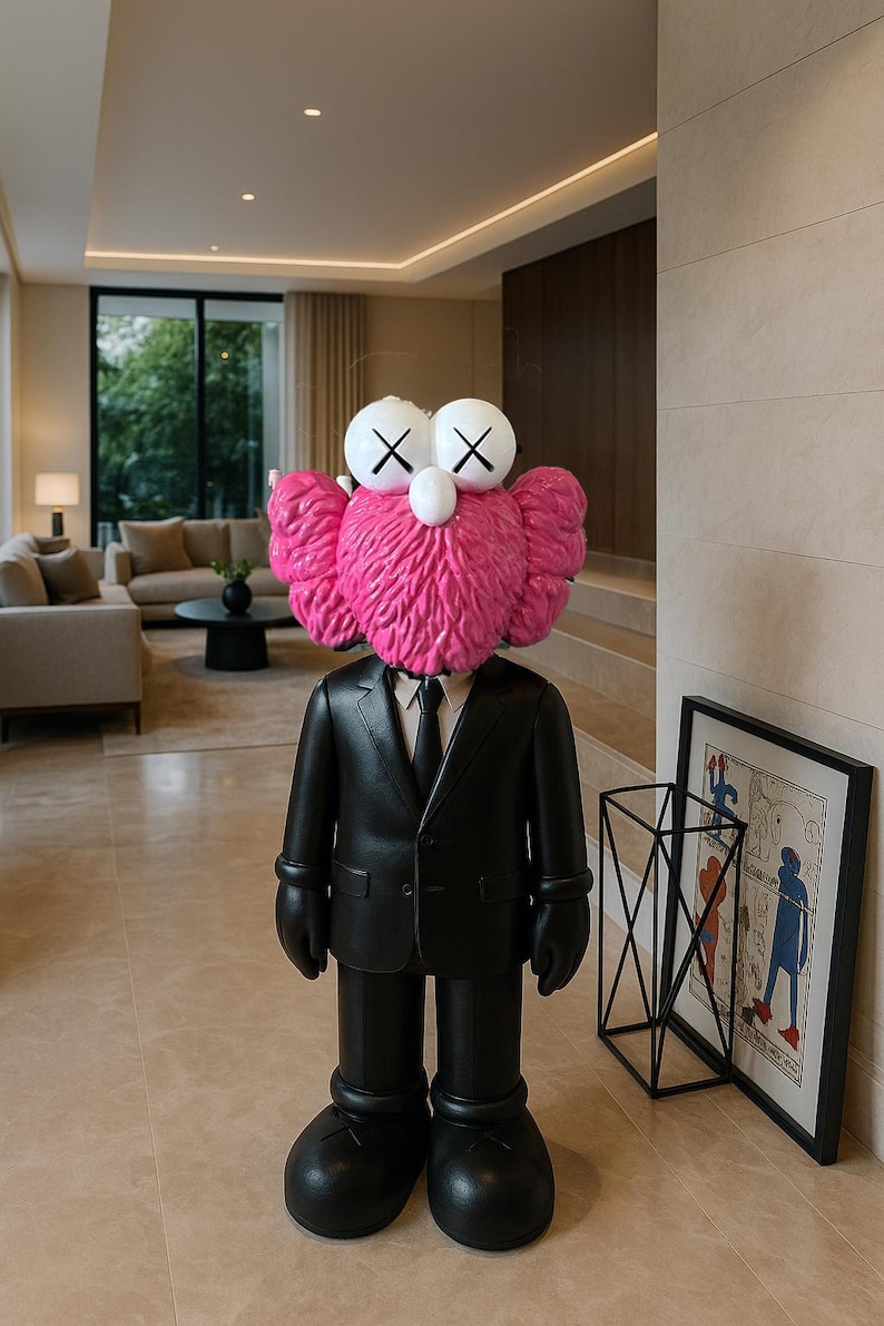 May include: A large, black sculpture of a figure in a suit with a pink, furry head and white eyes with black X's. The figure is standing in a modern, well-lit living room. A framed artwork and a black metal geometric sculpture are visible.