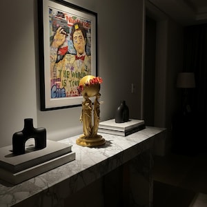 Könnte beinhalten: Ein gerahmtes Kunstwerk mit einem Porträt und Text im Graffiti-Stil hängt über einer Marmor-Konsole. Auf dem Tisch befindet sich eine goldfarbene Skulptur von Figuren, die einen Globus halten, zusammen mit einer schwarzen Vase und einer schwarzen abstrakten Skulptur.