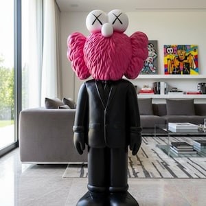 May include: A large black sculpture of a cartoon figure wearing a suit, with a pink head and ears, and white eyes with black X's. The figure is standing in a modern living room with large windows and artwork.