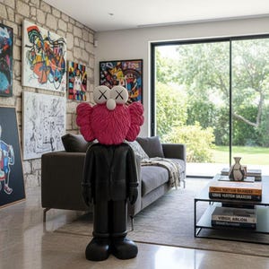 May include: A black and pink KAWS Companion figure stands in a modern living room with artwork on the stone wall. The figure has a pink head and a black suit. A gray sofa and coffee table with books are also visible.