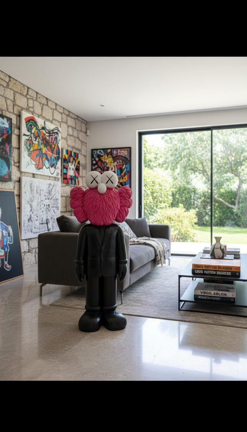 May include: A black and pink KAWS Companion figure stands in a modern living room with artwork on the stone wall. The figure has a pink head and a black suit. A gray sofa and coffee table with books are also visible.