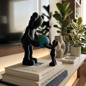 May include: Black sculpture of two figures holding a globe, placed on a stack of books. The larger figure is passing the globe to the smaller one. The books have titles like "The World as Art" and "Curated Living".
