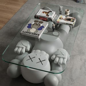 Pode incluir: Uma mesa de centro moderna com uma grande figura cinzenta em estilo cartoon e um tampo de vidro. A figura tem olhos riscados e m&atilde;os. Livros e pequenas figuras s&atilde;o exibidos na superf&iacute;cie de vidro. Uma pe&ccedil;a de mobili&aacute;rio &uacute;nica.
