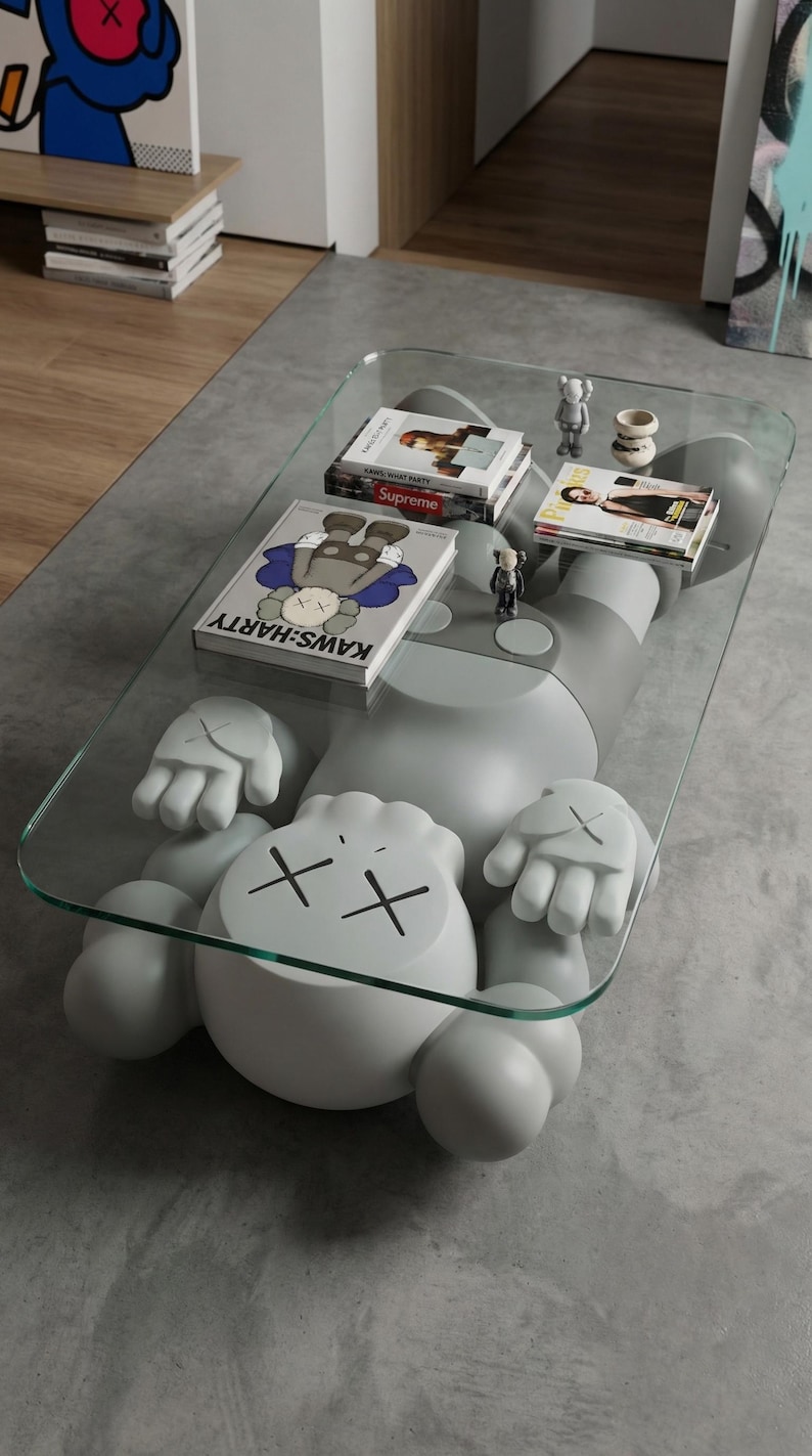 Pode incluir: Uma mesa de centro moderna com uma grande figura cinzenta em estilo cartoon e um tampo de vidro. A figura tem olhos riscados e m&atilde;os. Livros e pequenas figuras s&atilde;o exibidos na superf&iacute;cie de vidro. Uma pe&ccedil;a de mobili&aacute;rio &uacute;nica.