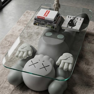 Pode incluir: Uma mesa de centro cinza &uacute;nica com um tampo de vidro transparente, em forma de personagem de desenho animado. A mesa tem m&atilde;os e p&eacute;s brancos, e o rosto do personagem apresenta olhos riscados. Livros e uma revista repousam sobre a superf&iacute;cie de vidro.