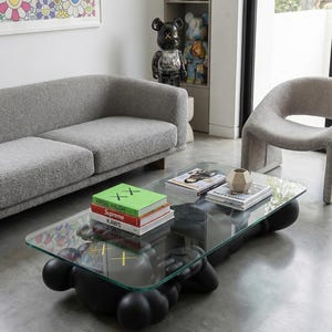 Pode incluir: Sala de estar moderna com uma mesa de centro com tampo de vidro sobre uma base preta, um sof&aacute; cinza e uma cadeira curva. Livros e objetos decorativos est&atilde;o sobre a mesa. Uma obra de arte floral est&aacute; pendurada na parede.