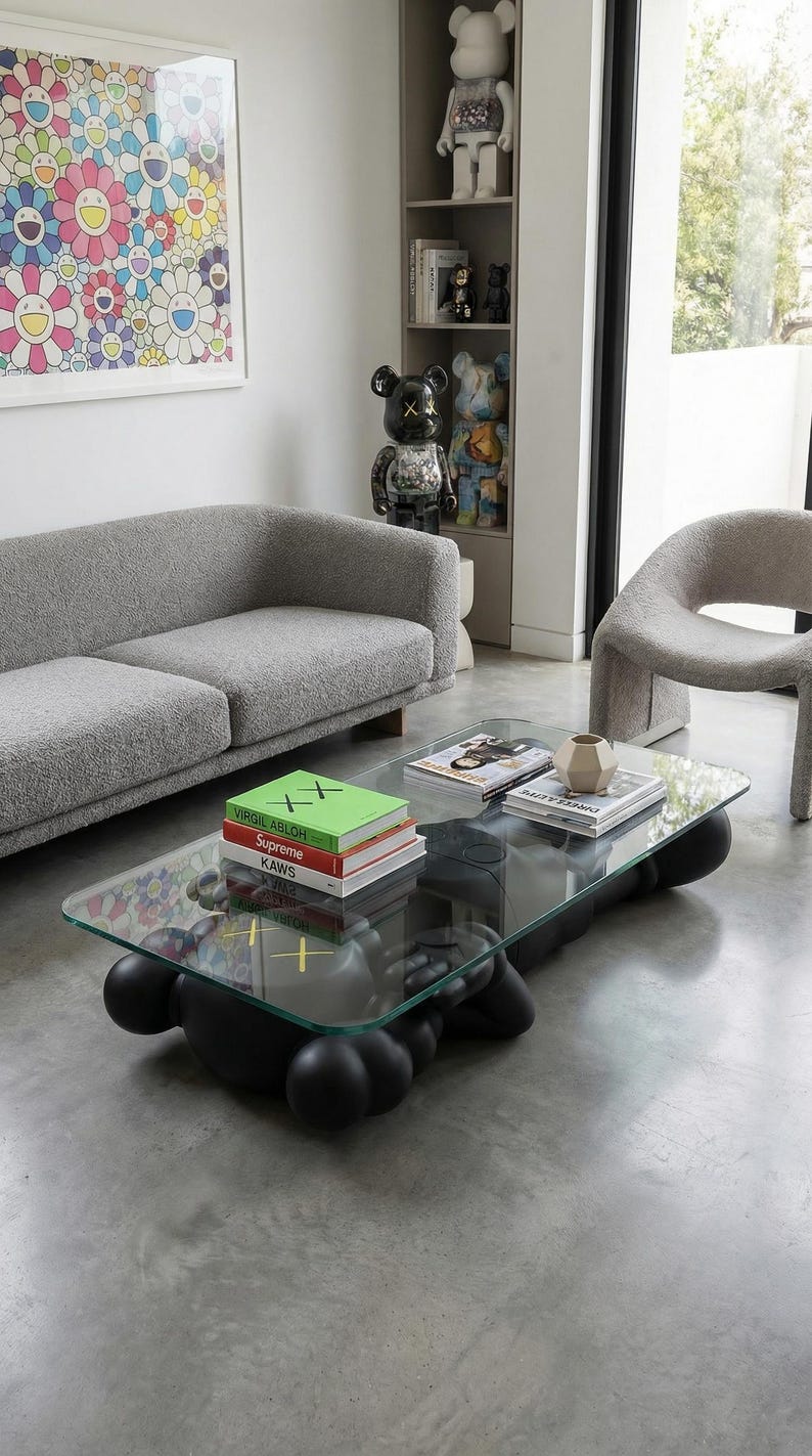 Pode incluir: Sala de estar moderna com uma mesa de centro com tampo de vidro sobre uma base preta, um sof&aacute; cinza e uma cadeira curva. Livros e objetos decorativos est&atilde;o sobre a mesa. Uma obra de arte floral est&aacute; pendurada na parede.