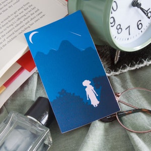 Puede incluir: Un marcapáginas ilustrado azul y blanco con una silueta de una persona de pie frente a una cordillera de montañas bajo una luna creciente y una estrella fugaz. El marcapáginas está en una cama con un libro, gafas y una botella de perfume.