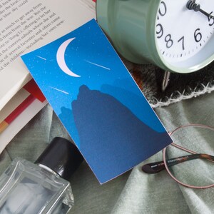Puede incluir: Una tarjeta ilustrada azul y blanca con una luna creciente y estrellas fugaces sobre una cordillera oscura. La tarjeta está sobre una manta verde y marrón con un libro, una botella de perfume y unas gafas.