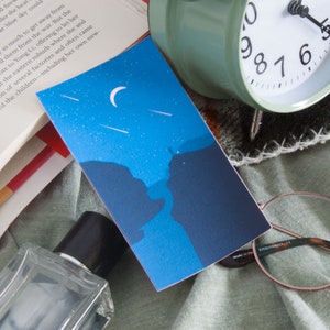 Puede incluir: Una tarjeta ilustrada azul y negra con una luna creciente y estrellas fugaces en el cielo nocturno. La tarjeta presenta una silueta de un acantilado con una persona de pie en la cima.