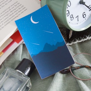 Puede incluir: Una tarjeta azul con una luna creciente y estrellas fugaces en el cielo nocturno sobre una cordillera azul oscuro. La tarjeta está sobre una superficie de tela verde y marrón con un libro, un reloj y gafas.